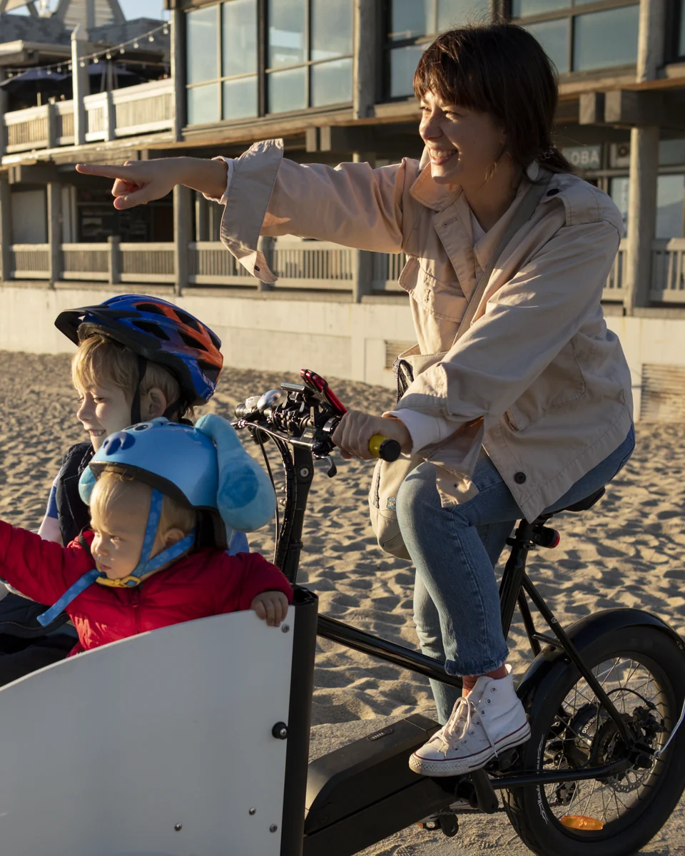 A mother riding an electric bike with two children on a beach path